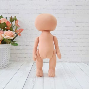 May include: A nude-colored fabric doll with a large head and limbs. The doll is standing on a white wooden surface.