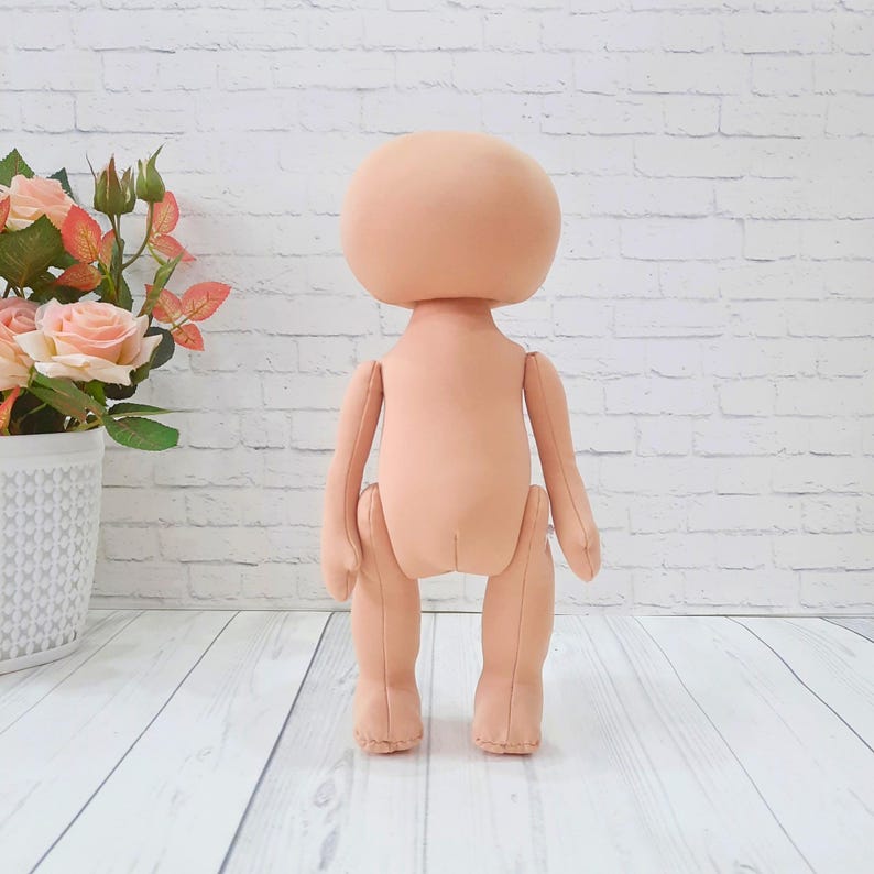 May include: A nude-colored fabric doll with a large head and limbs. The doll is standing on a white wooden surface.
