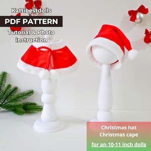 Op de afbeelding: Een rode en witte kerst hoed en cape voor poppen van 25-28 cm. De hoed heeft een witte rand en een witte pompon. De cape heeft een witte bontkraag en twee witte pompoenen.