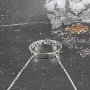 Può includere: Un ciondolo a forma di anello in argento con l'iscrizione "SAM & LIAM" sull'anello. L'anello è attaccato a una catena d'argento.