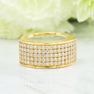 Könnte beinhalten: Ein breiter, goldfarbener Ring mit mehreren Reihen kleiner, funkelnder Diamanten. Das Band des Rings ist dick und weist ein durchgehendes Muster der Edelsteine auf, wodurch ein brillantes Spiel von Licht und Farbe entsteht. Der Ring ist vor einem weichen, verschwommenen Hintergrund platziert.