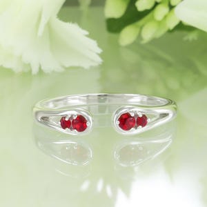 May include: A silver ring with three bright red gemstones set in each teardrop-shaped end. The ring has an open design, with the gemstones visible from the top. The ring is displayed on a reflective surface.