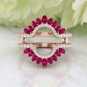 May include: A rose gold ring guard with a double band design. The bands are adorned with a row of small, clear gemstones and a series of vibrant, teardrop-shaped, red gemstones, creating a sunburst effect. The ring guard is designed to enhance the look of a solitaire ring.