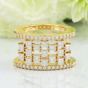 Könnte beinhalten: Ein goldener Ring mit einem einzigartigen Design. Der Ring zeigt eine Reihe quadratisch geschliffener Diamanten in einem Gittermuster, mit kleineren Diamanten, die die oberen und unteren Bänder umgeben. Der Ring liegt auf einer weißen Oberfläche.