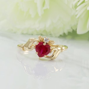 以下が含まれることがあります： ハート型の赤い宝石が付いたゴールドカラーのリング。リングのデザインは、ハートを抱える2つの手と、その上にある小さなダイヤモンドと葉のアクセントが特徴です。背景は柔らかくぼやけており、リングの詳細を際立たせています。