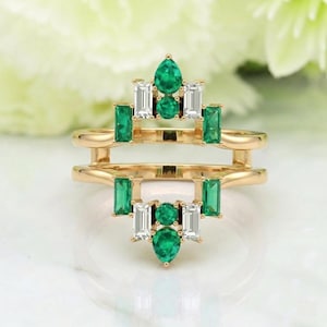 May include: A gold-colored ring with emerald and white gemstone accents. The ring features a unique design with two bands connected by vertical supports. The gemstones are emerald green and white, arranged in geometric patterns.