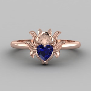 May include: Rose gold ring with a fox motif and a heart-shaped, deep blue gemstone. The fox's head and ears are at the top, with the gemstone at the centre. The ring band is smooth and rounded.