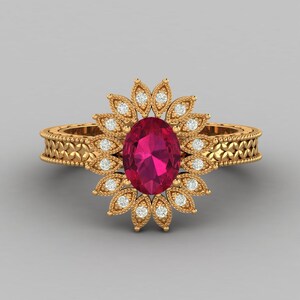 Op de afbeelding: Gouden ring met een grote, ovaal geslepen, robijnrode edelsteen in het midden, omringd door kleine, heldere edelstenen. De ringband heeft een gedetailleerd bloemenpatroon.