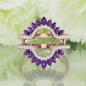 Puede incluir: Un protector de anillo de oro rosa con un diseño de doble banda. Cada banda presenta una fila de pequeños diamantes blancos y una fila curva de piedras preciosas moradas en forma de lágrima. El protector de anillo está diseñado para realzar el aspecto de un anillo de compromiso.