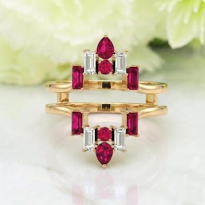 May include: A gold-colored ring with a double band design. The ring is adorned with a series of red and white gemstones. The gemstones are arranged in a symmetrical pattern, with pear-shaped, rectangular, and round stones.