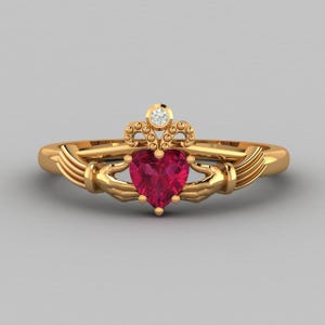 May include: A gold-colored Claddagh ring featuring a heart-shaped red gemstone, a small diamond, and hands holding a crown. The ring's design is a symbol of love, loyalty, and friendship.