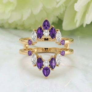 Puede incluir: Un anillo dorado con un diseño de doble banda. El anillo está adornado con una variedad de piedras preciosas, incluyendo piedras blancas en forma de marquesa, piedras moradas en forma de pera y redondas. El fondo es suave y borroso.