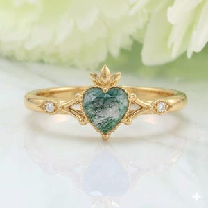 May include: A gold ring featuring a heart-shaped green moss agate gemstone. The ring has a delicate design with small diamonds on each side of the heart and a crown-like detail above the heart. The ring is set on a gold band.