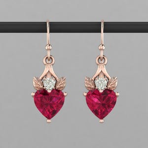 May include: A pair of rose gold earrings. Each earring features a heart-shaped, deep red gemstone, accented with a small, round, clear stone and leaf-shaped details. The earrings hang from a simple hook.