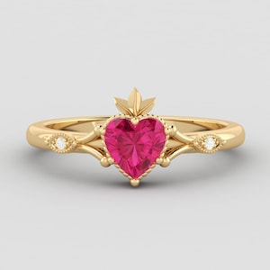 Pode incluir: Um anel de ouro com uma pedra preciosa rosa em forma de coração e facetada. O anel tem um design decorativo com pequenos diamantes e um detalhe em forma de folha acima do coração. A banda é lisa e polida.
