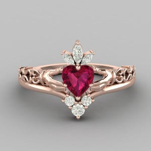 Peut inclure: Bague Claddagh en or rose avec un rubis en forme de cœur, encadré de mains et surmonté d'une couronne de diamants. La bague présente des détails de filigrane complexes sur le bandeau.