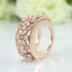 Könnte beinhalten: Roségoldener Ring mit floralem Design. Der Ring ist mit zahlreichen klaren, funkelnden Edelsteinen in verschiedenen Formen besetzt, die ein schillerndes Schauspiel erzeugen. Das Band hat eine glatte, polierte Oberfläche.