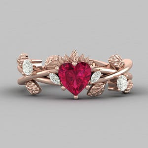 Pode incluir: Anel em ouro rosa com uma pedra vermelha em forma de coração no centro, rodeada por detalhes de folhas e videiras. Acentuado com pequenas pedras brancas redondas. Um design romântico e inspirado na natureza.