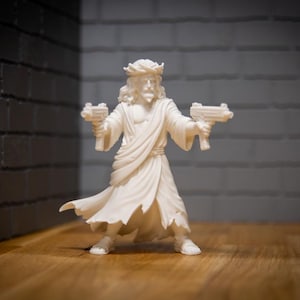 May include: A white figurine of a figure with long hair and a crown, holding two toy guns. The figure is wearing a robe and standing on a wooden surface. The background features a brick wall and a dark grey wall.