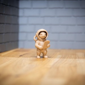 May include: A tan-coloured figurine of a frog in a spacesuit, holding a clipboard. The frog is wearing a helmet with a hose and boots. The figurine is standing on a wooden surface.