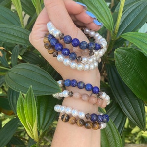 May include: Several beaded bracelets in various styles. One bracelet features white pearls, another has blue lapis lazuli beads, and others have tiger's eye and peach-colored beads. The bracelets are stacked on a wrist against a backdrop of green foliage.