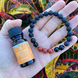 May include: Aromatherapy essential oils bottle with an orange label next to a black lava stone bracelet with black, red, and gold beads. The bracelet is held in a hand.