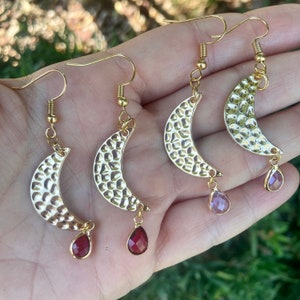 May include: Gold-toned crescent moon earrings with a hammered texture. Each earring features a teardrop-shaped gemstone pendant in either a deep red or a light purple hue. The earrings have a hook closure.