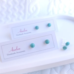 Może przedstawiać: Turkusowe kolczyki sztyfty prezentowane na białej tacy z napisem "Azalea Handmade Accessories" na białej karcie. Widoczne są również luźne turkusowe koraliki. Kolczyki są małe, okrągłe i w żywym turkusowym kolorze.