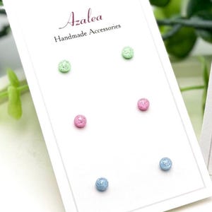 May include: A set of three pairs of stud earrings in pastel colors: green, pink, and blue. Each pair is displayed on a white card with the text "Azalea Handmade Accessories". The earrings are presented on a white, oval tray with green foliage in the background.