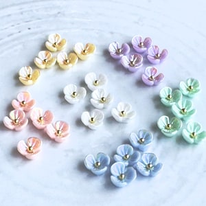 May include: A collection of small, flower-shaped embellishments in various pastel colors, including yellow, pink, white, purple, blue, and green. Each flower has a gold-colored center. The arrangement is on a light-colored, textured surface, suitable for crafts or jewelry making.