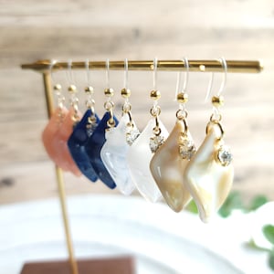 May include: A collection of dangle earrings in various colors, including pink, navy blue, white, and beige. Each earring features a diamond-shaped pendant with a small, clear gemstone and gold-colored accents. The earrings are displayed on a gold-colored stand.