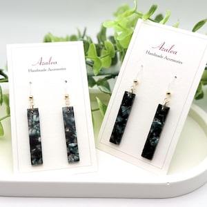 May include: Two pairs of Azalea Handmade Accessories earrings. Each earring features a rectangular, marbled black and teal design with a gold-colored bead and clear hook. The earrings are displayed on white cards.