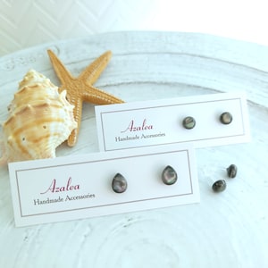 May include: Two pairs of Azalea Handmade Accessories earrings. One pair is round, the other is teardrop-shaped. The earrings are dark with iridescent coloring. The earrings are displayed on white cards with the brand name and a starfish and seashell in the background.