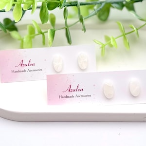 May include: Two pairs of white oval-shaped stud earrings on display cards. The cards are pink and white with the word "Azalea" and "Handmade Accessories" printed on them. The earrings are on a white tray with green foliage.