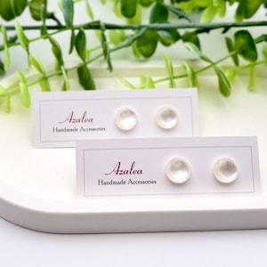 May include: Two pairs of white, round stud earrings displayed on cards that read "Azalea Handmade Accessories." The cards are on a white, oval tray, with green foliage in the background.