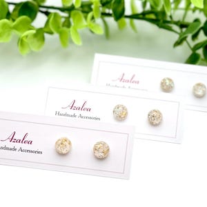 以下が含まれることがあります： キラキラとした質感の丸いスタッドピアスが3組、白いカードに展示されています。カードには筆記体で「Azalea Handmade Accessories」と書かれています。背景には緑の葉があります。