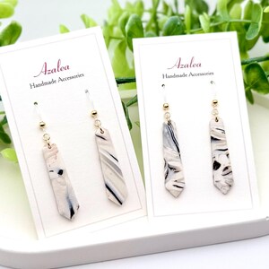 May include: A pair of white and black marble-patterned earrings with gold hooks. The earrings are displayed on white cards with the text "Azalea Handmade Accessories".
