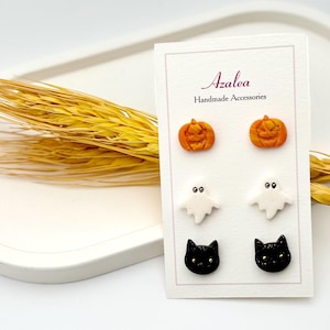 Op de afbeelding: Een kaart met zes Halloween-thema oorknopjes. De oorbellen hebben oranje pompoenen, witte geesten en zwarte katten. De kaart leest "Azalea Handmade Accessories". De oorbellen staan op een witte achtergrond met tarwearen.