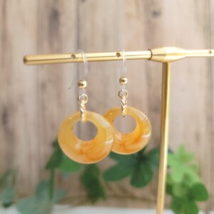 May include: A pair of amber-coloured dangle earrings with a circular design. The earrings have a translucent, marbled effect. They are suspended from gold-coloured hooks and small gold beads. The earrings are displayed on a gold-coloured jewellery stand.