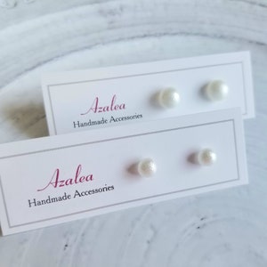 Op de afbeelding: Twee paar witte parelstud oorbellen gepresenteerd op witte kaarten. De kaarten hebben een grijze rand en het woord "Azalea" in een cursief lettertype, met "Handmade Accessories" eronder. De oorbellen zijn rond en hebben een subtiele glans.