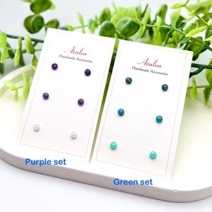 Puede incluir: Dos juegos de pendientes en tarjetas blancas. La tarjeta de la izquierda tiene un juego de pendientes morados, mientras que la tarjeta de la derecha tiene un juego de pendientes verdes. Ambas tarjetas tienen el texto "Azalea Handmade Accessories" y las palabras "Purple set" y "Green set" en la parte inferior.