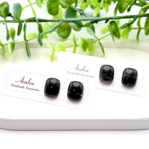 May include: Black and gray square stud earrings on white cards with the name "Azalea" and "Handmade Accessories" printed in a script font. The earrings are displayed on a white oval tray with green foliage.