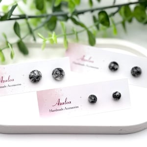 May include: A display of Azalea Handmade Accessories earrings. The earrings are small, round studs with a marbled black and white pattern. The earrings are displayed on white cards with the Azalea logo and text.