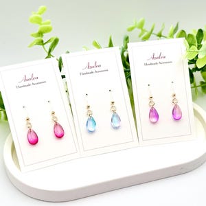 May include: Three pairs of Azalea handmade earrings on display. Each pair features a teardrop-shaped charm in shades of pink, blue, and purple. The earrings are on white cards with the Azalea logo and are displayed on a white tray.