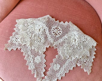Puede incluir: Un cuello de encaje antiguo, de color blanquecino, con diseños florales y circulares. El cuello está colocado sobre una silla rosa tapizada. El encaje tiene un borde festoneado y probablemente está hecho a mano.