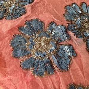 May include: Close-up of a coral pink fabric with a repeating pattern of large, gray and gold floral embellishments. The embellishments are made of sequins and beads.