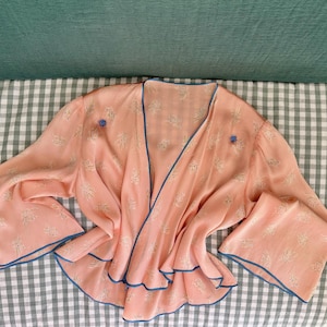 Könnte beinhalten: Eine pfirsichfarbene Wickelbluse mit zartem weißem Blumenmuster und kontrastierender blauer Paspelierung entlang des V-Ausschnitts, des Saums und der weiten Ärmel. Das Kleidungsstück hat zwei kleine blaue kreisförmige Akzente im Brustbereich.