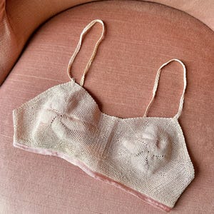 Può includere: Un bralette all'uncinetto color beige caratterizzato da due motivi circolari ispirati ai fiori sulle coppe, sottili spalline intrecciate e una fascia liscia in tinta unita lungo il bordo inferiore. Il capo è realizzato con un punto a rete fine.