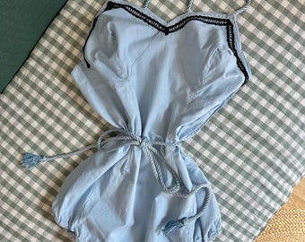 May include: A light blue romper featuring thin braided rope straps and a matching waist tie with tassel ends. The bodice includes a black decorative trim along the neckline, while the bottom portion has gathered leg openings for a bubble silhouette.