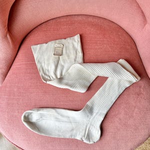May include: A pair of white knee-high socks with a ribbed texture. The socks are lying on a pink chair. A small white tag with the text "Symphonie" is attached to the socks.